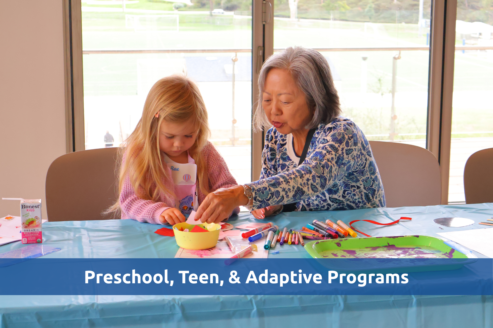 Adult helps young child with a craft activity at a table with markers and art supplies.